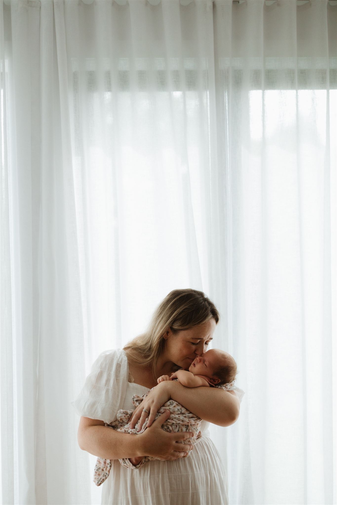 Mother holding baby against sheer curtains and kissing her gently.