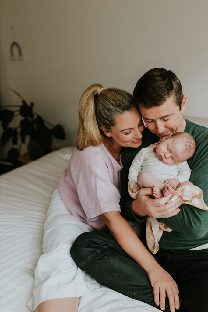 Parents sitting on the end of their bed cuddling their newborn baby girl.
