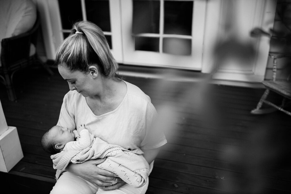 Black and white image of mother sitting on porch holding sleeping baby.