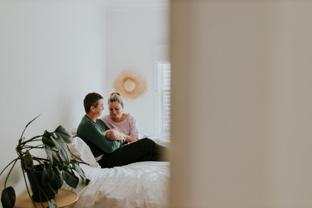 Parents sitting on their bed looking and smiling at their newborn baby.
