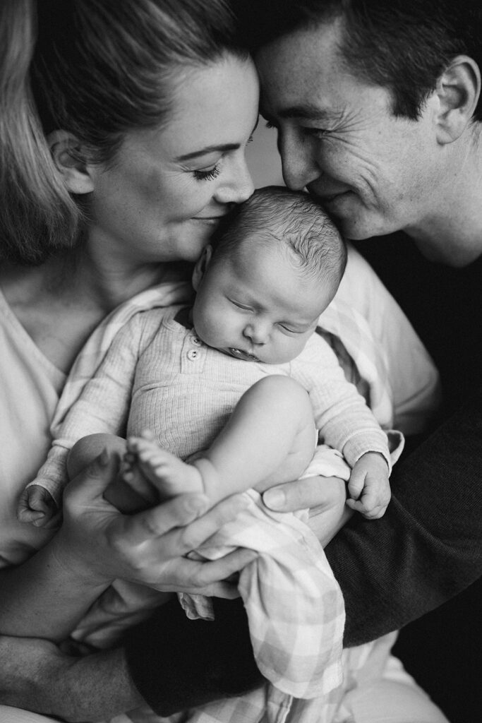 Close up of parents faces kissing their newborns baby's head who is asleep.