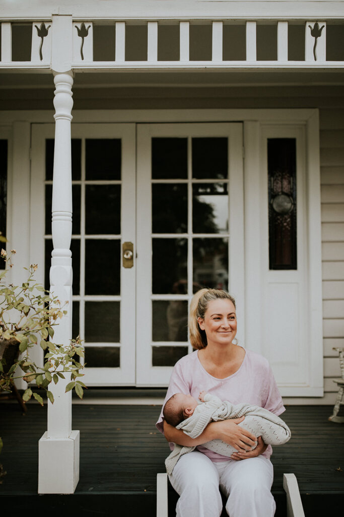 Smiling mother sitting outside on her porch holding her newborn baby who is wrapped in a blanket. 