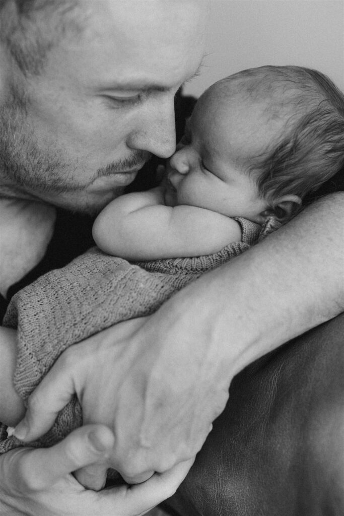Dad and newborn baby touching noses while baby sleeps in his arms.
